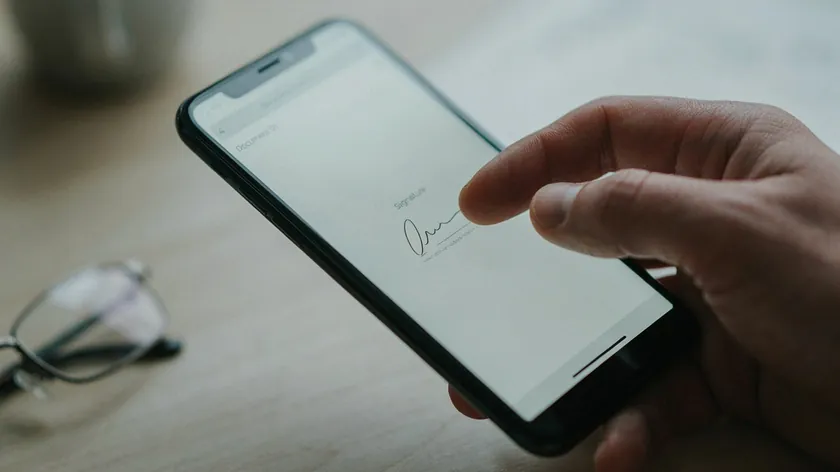 Finger drawing a handwritten signature on an iPhone screen over a blank document